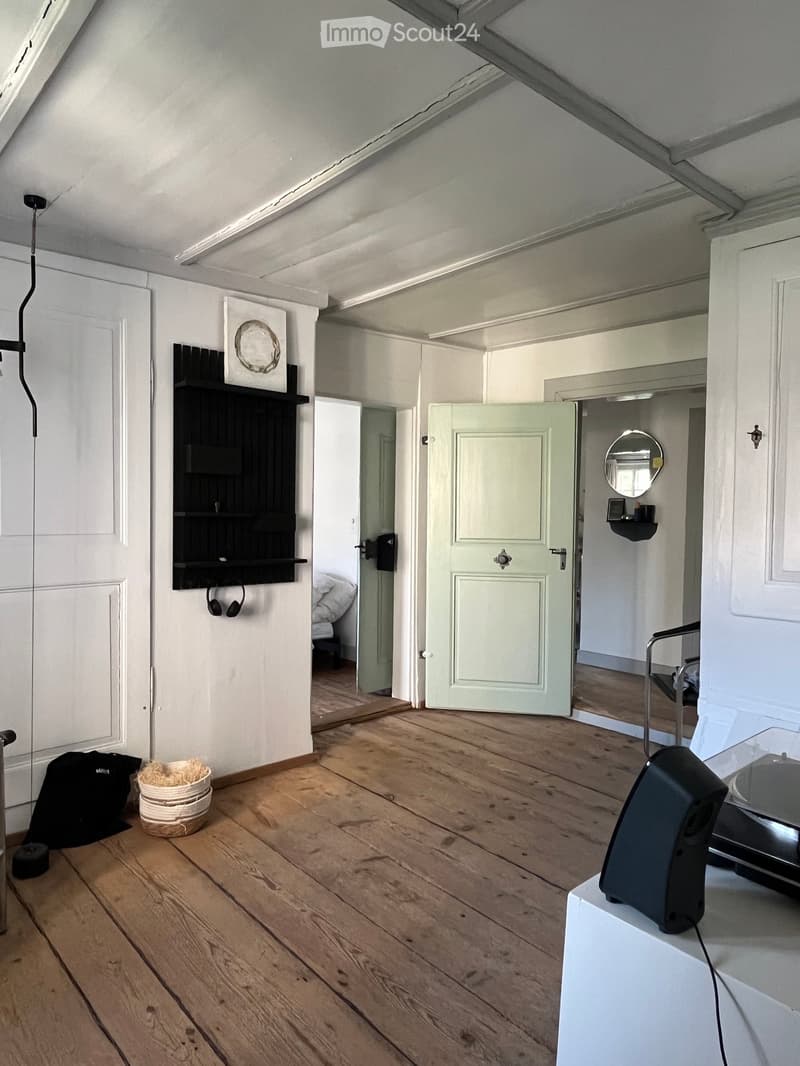 A modern, minimalist room with wooden flooring, white walls, and a black shelf with headphones. A speaker is placed on a white surface, and a mirror is visible in an adjacent room.
