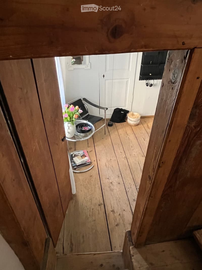 Narrow wooden hallway with a small round table, chair, vase with flowers, magazines, and a door leading to another room.