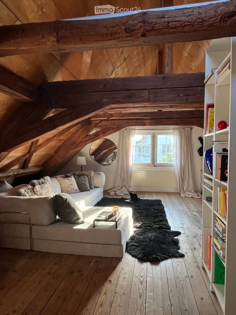 Cozy attic room with exposed wooden beams, large windows, a white sofa with pillows, a coffee table, a bookshelf, and a fur rug on the floor.