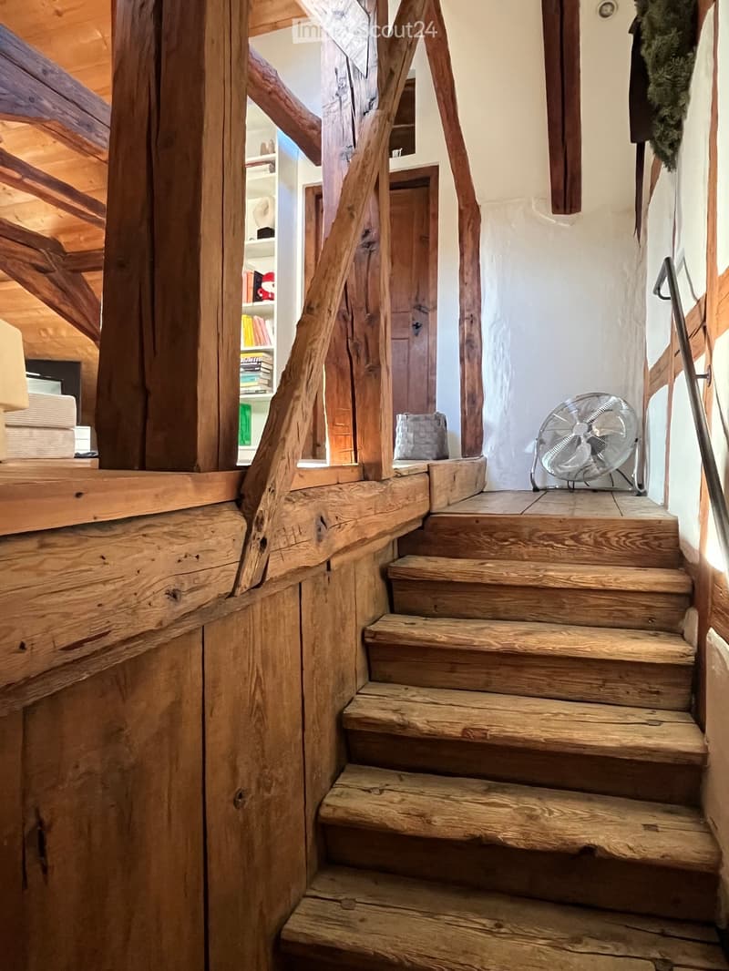Wooden staircase with hand-carved balusters and a metal railing, leading to an open area with a bookshelf and a fan on the landing.