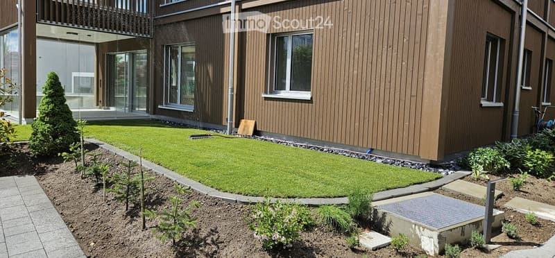 Garden / Outdoor Area. Well-maintained garden with a neatly trimmed lawn, small trees, shrubs, and a paved pathway leading to a modern building with large windows and a balcony.