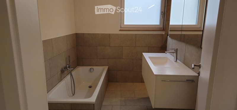 Bathroom. Modern bathroom with a bathtub, sink, and large mirror. The walls are tiled in neutral tones, and the space is well-lit with natural light from the windows.
