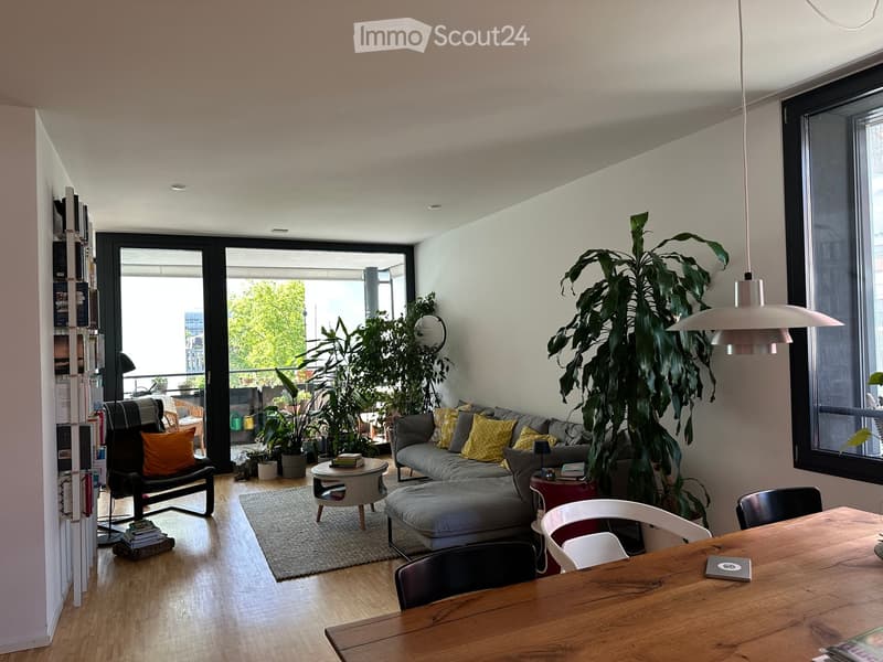 Modern living room with a grey sectional sofa, coffee table, dining table, chairs, bookshelf, and large plants. The space features large windows and a sliding glass door leading to a balcony.
