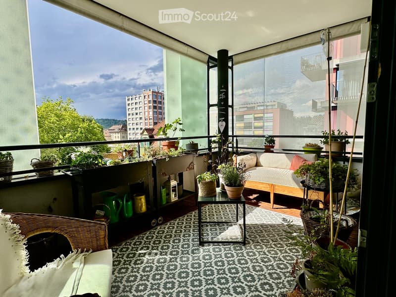 Balcony with a sofa, small table, chair, and multiple potted plants, overlooking a cityscape with buildings and trees.