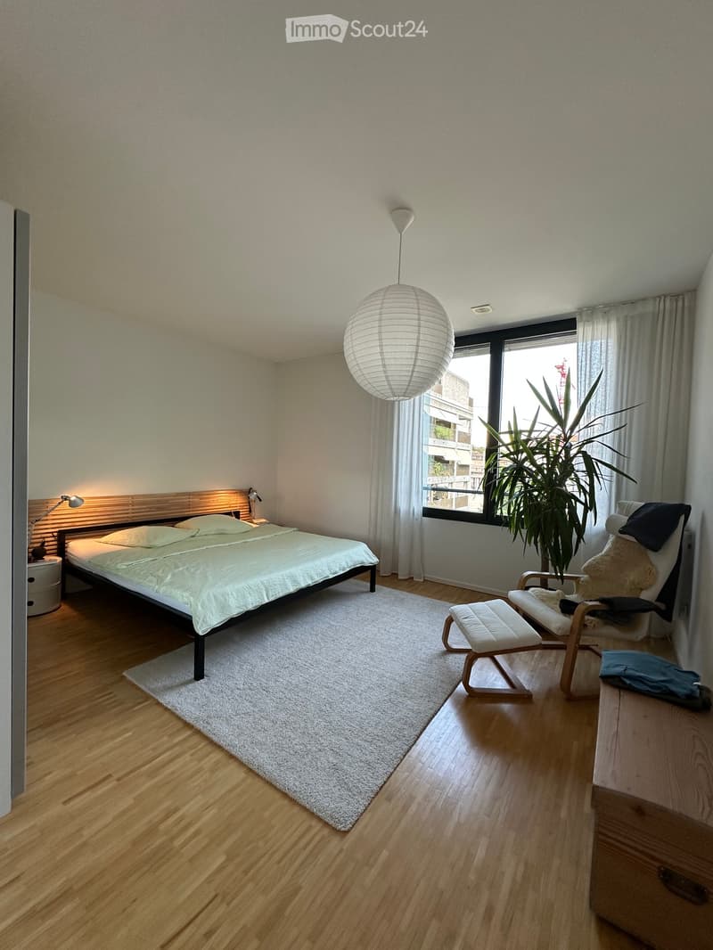 Modern bedroom with a large bed, white bedding, a hanging spherical lamp, a wooden chair with a cushion, a small stool, a potted plant, and a nightstand with a lamp.
