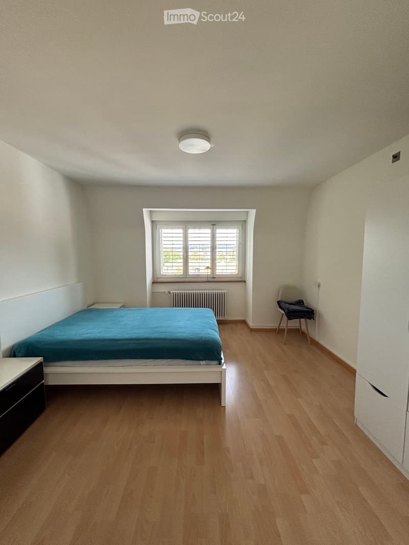A furnished bedroom with a wooden floor, a single bed with blue bedding, a nightstand, a chair, and a radiator beneath a window.