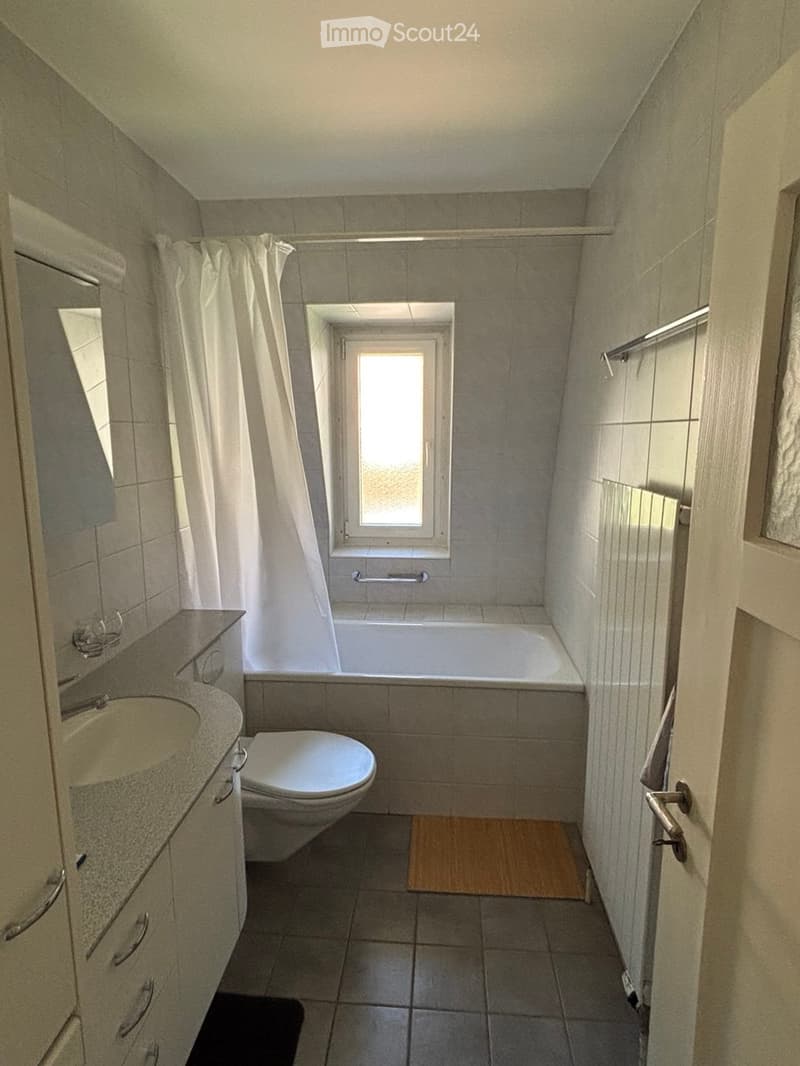 Modern bathroom with a bathtub, toilet, sink, and mirror. Features a window above the bathtub and a wooden bath mat on the floor.