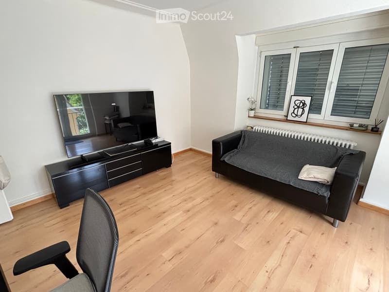Modern living room with light wooden flooring, a black leather sofa, a TV stand with a large flat-screen TV, a black chair, and a mirror on the wall.