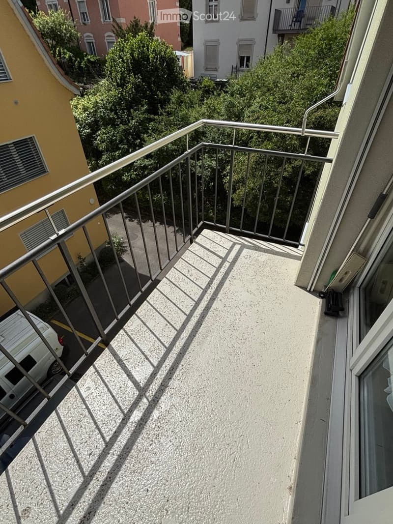 Balcony with metal railing, concrete floor, and view of neighboring buildings and greenery.