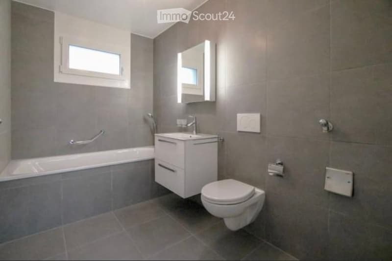 Modern bathroom with grey tiled walls and floor, featuring a bathtub, a white sink with a mirror, and a toilet.