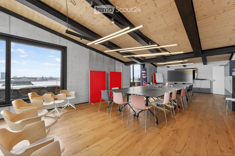 Modern office space with wooden flooring, large windows, and contemporary furniture including a long conference table with chairs, a large screen, and additional screens on the wall.