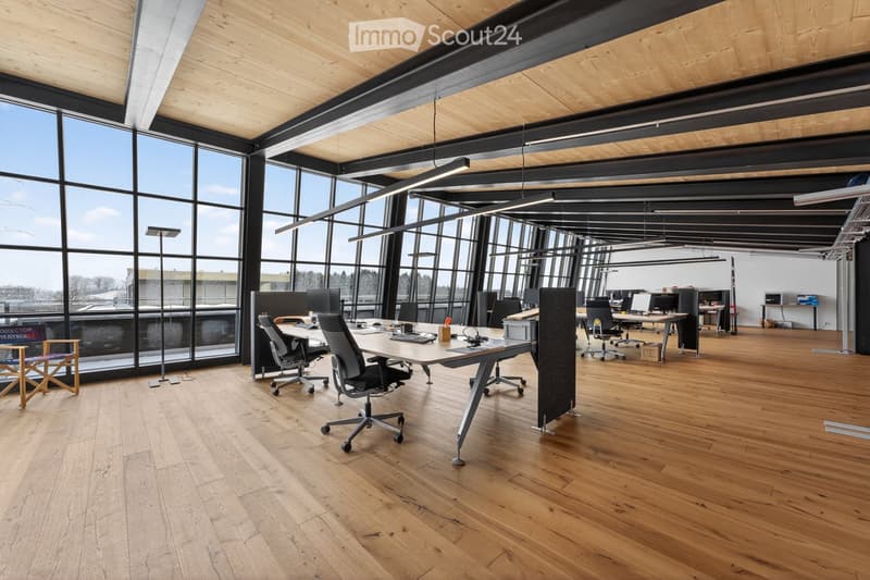 Modern office space with large windows, wooden flooring, and contemporary furniture including office tables, chairs, and computer monitors.