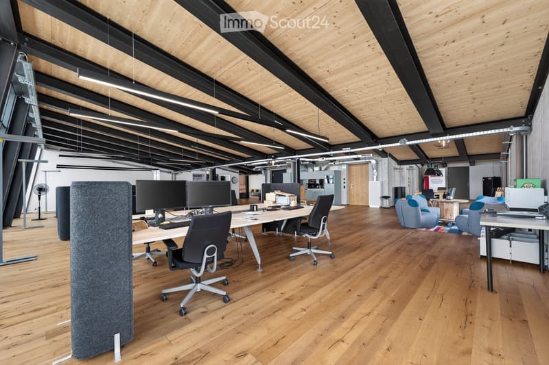 Modern office space with wooden flooring, exposed wooden beams, and black metal structural elements. Features office chairs, desks with computer monitors, keyboards, a screen divider, and a small seating area with a sofa and fan.