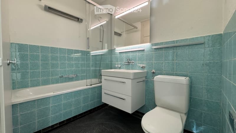Modern bathroom with teal tiled walls, white fixtures including a bathtub, sink, and toilet, and a glass-enclosed shower area.