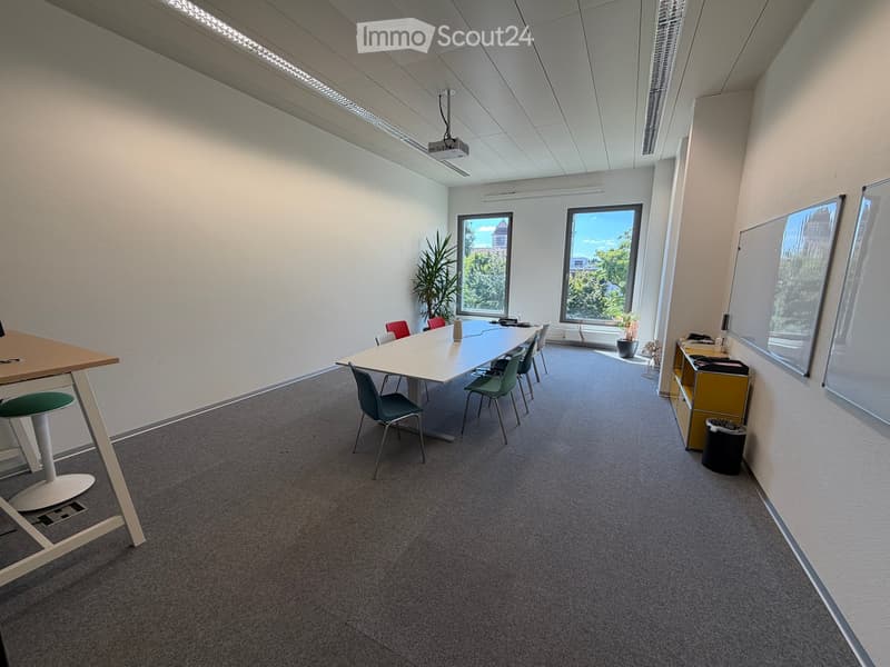 Bright, modern meeting room with a long table, chairs, a screen, and a small cabinet. Large windows provide natural light and a view of greenery.