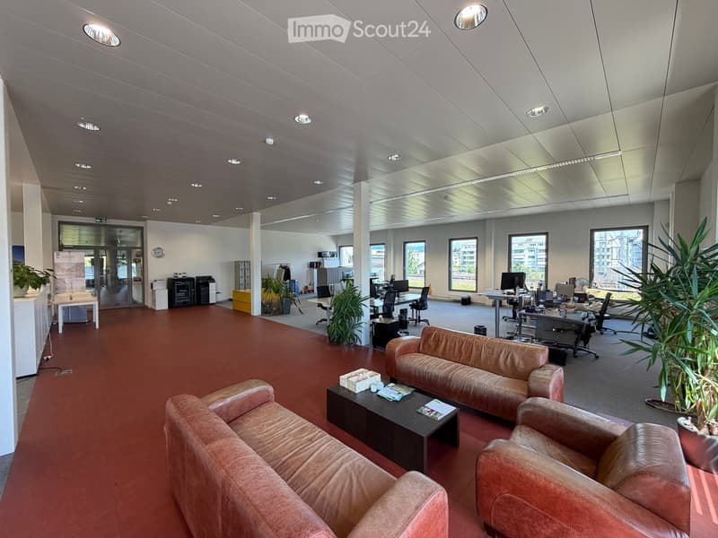 Modern office space with red carpeting, white walls, and a high ceiling. Features a leather sofa, coffee table, office chairs, desks, and large windows with a city view.