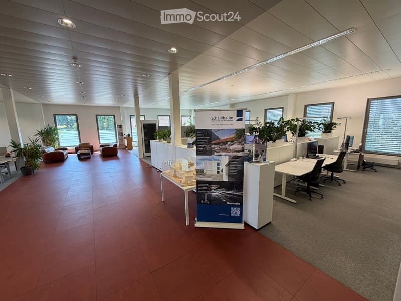 Office space with a mix of seating areas and workstations, featuring a red-tiled floor, large windows, and modern furniture.