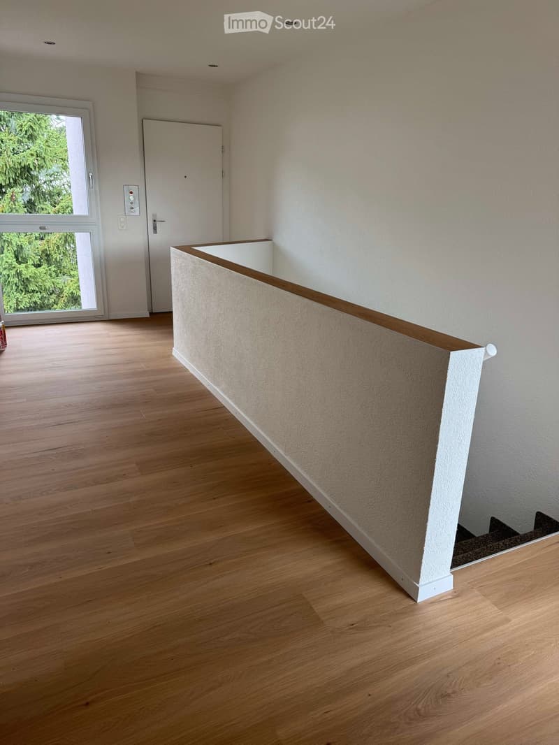 Empty room with wooden flooring, white walls, and a staircase leading upwards. A large window with a view of greenery is visible on the left side.