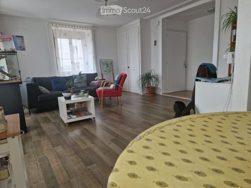 Living room with wooden flooring, a dark blue sofa, a red armchair, a small white side table, a fish tank, and a potted plant. The room has large windows with white curtains and a ceiling light fixture.