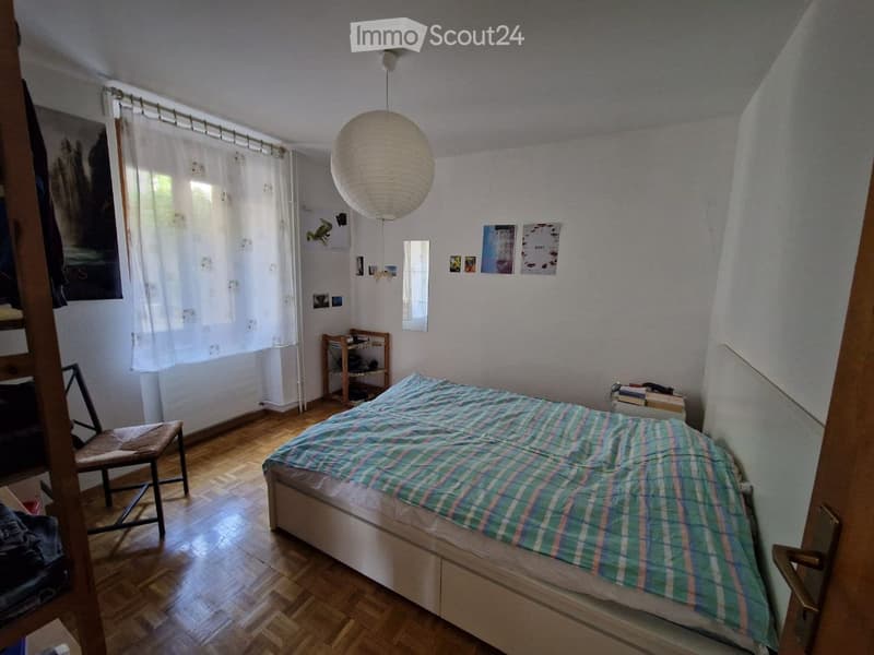Cozy bedroom with wooden flooring, a bed with a green and white checkered duvet, a wooden chair, a small shelf, and a hanging paper lantern light fixture.