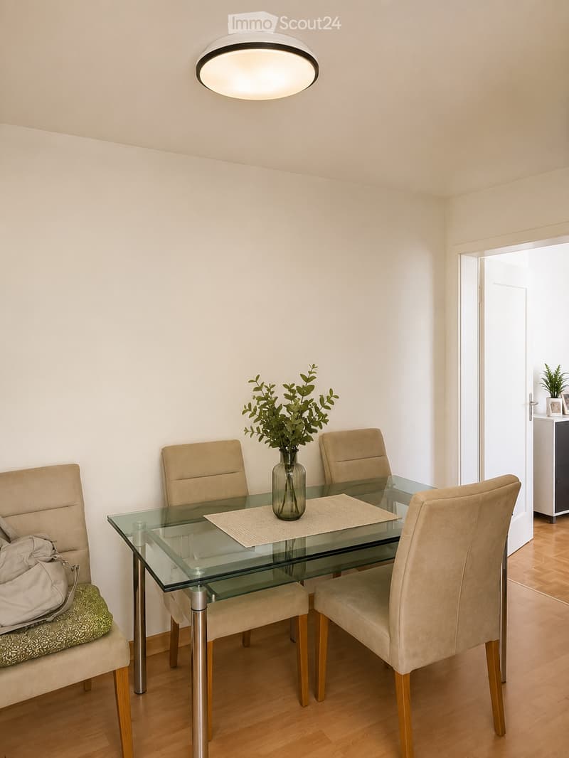 Dining room with a glass table, four beige chairs, a vase with greenery, and a handbag on a chair.