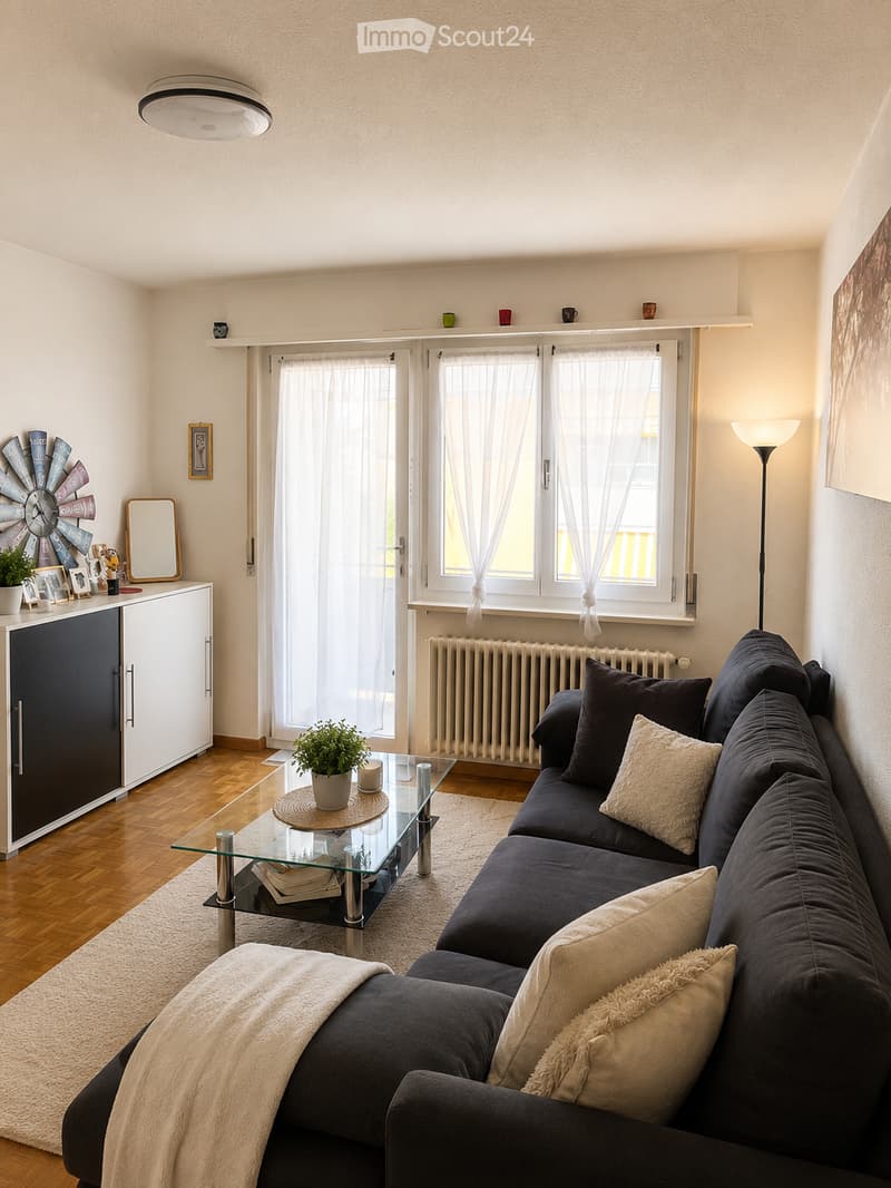 Modern living room with dark grey sectional sofa, glass coffee table, floor lamp, and a white cabinet with a mirror. The room features wooden flooring, large windows with white blinds, and a small plant on the coffee table.