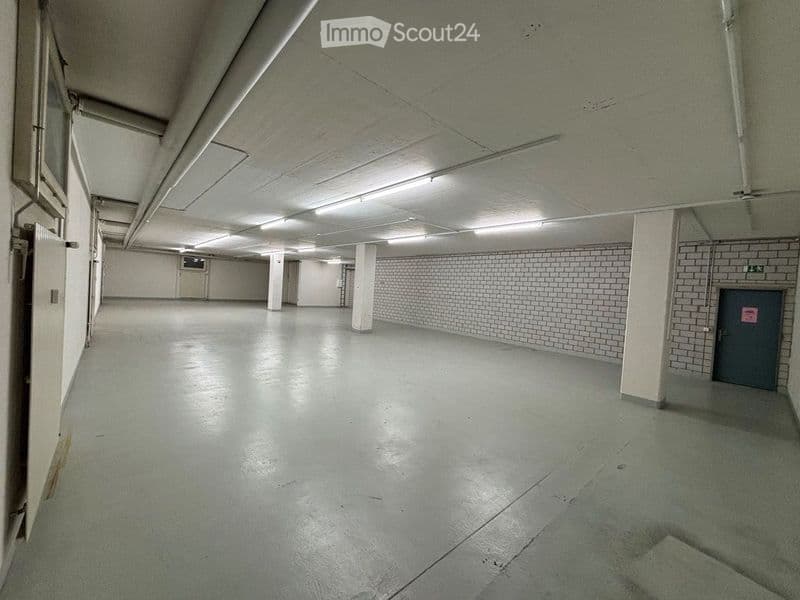 Empty, spacious room with concrete floors and white brick walls, illuminated by overhead fluorescent lighting. The room features several white support columns and a single grey door on the right side.