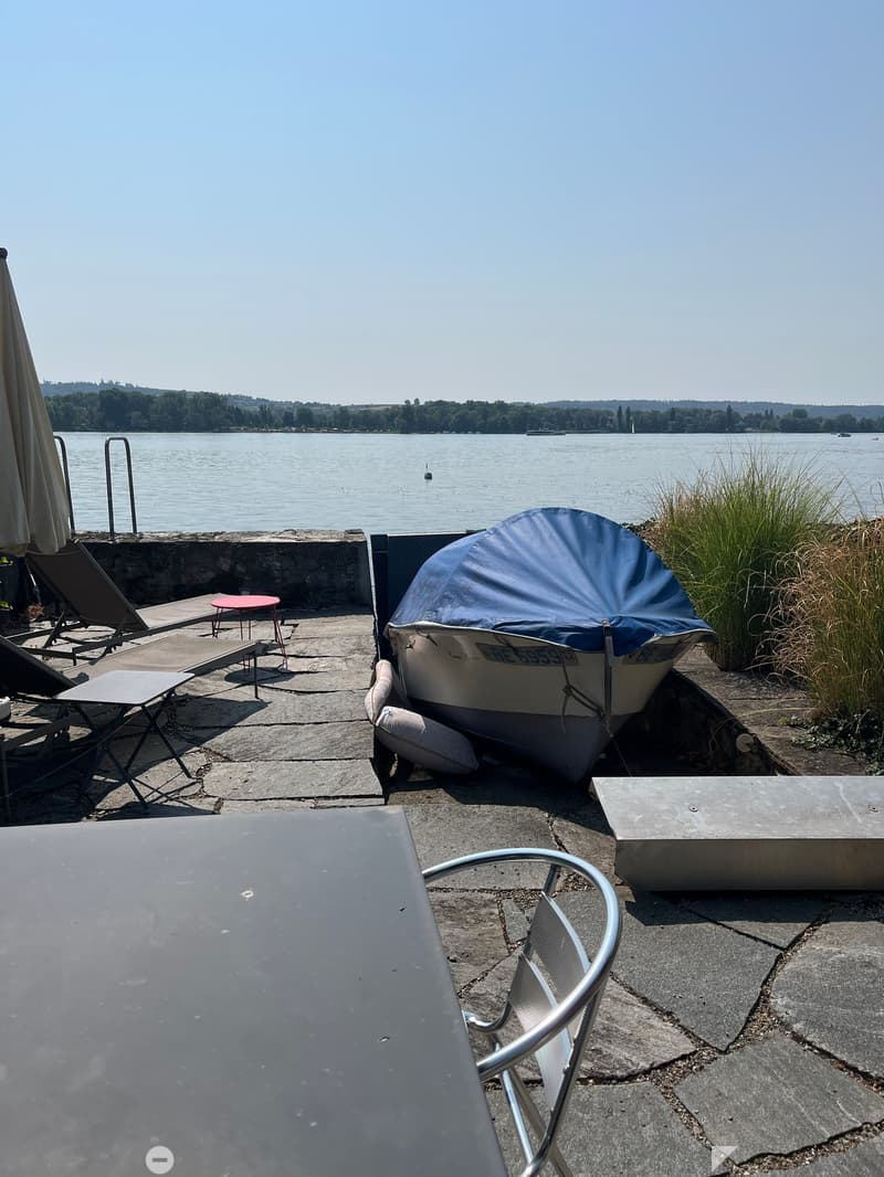 Outdoor area by a lake with a covered boat, tables, chairs, and an umbrella, overlooking the water.