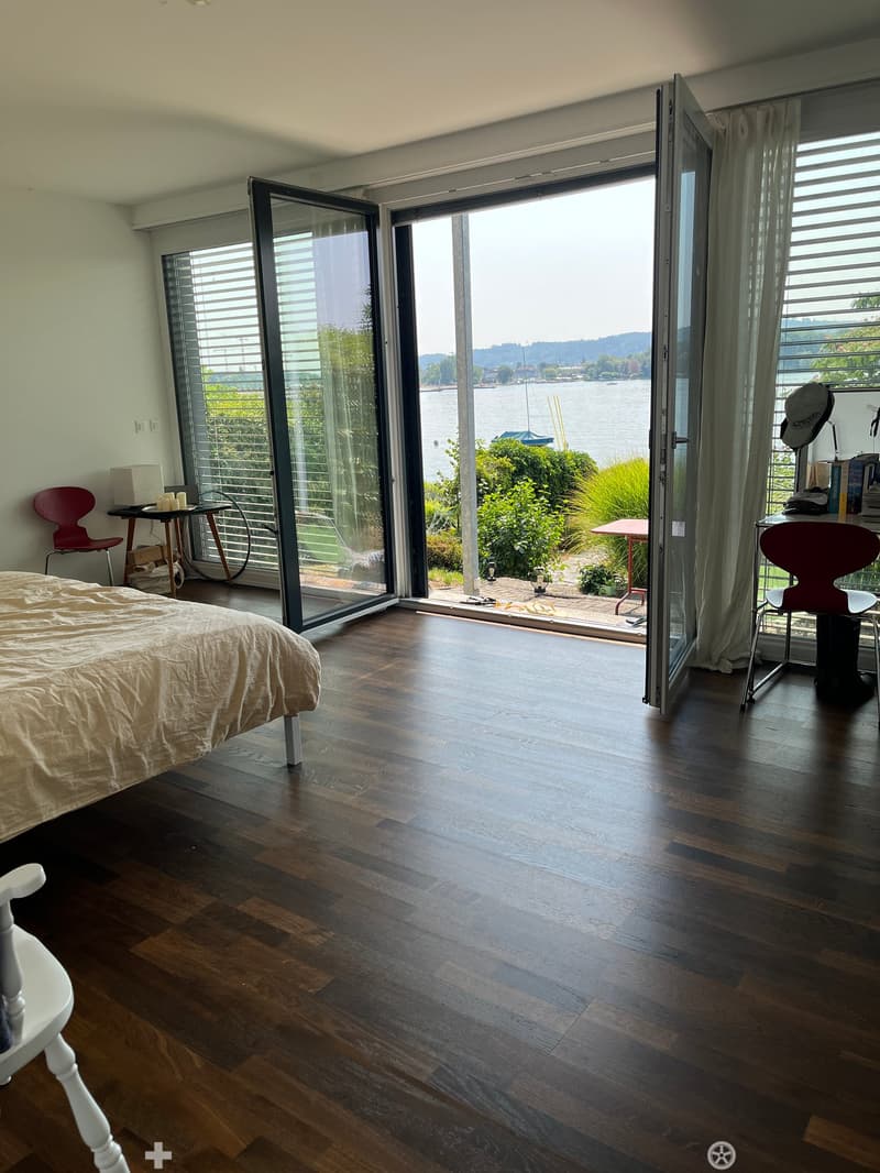 Bright bedroom with wooden flooring, large sliding glass doors opening to a scenic waterfront view, a bed, a red chair, a small table, and a lamp.