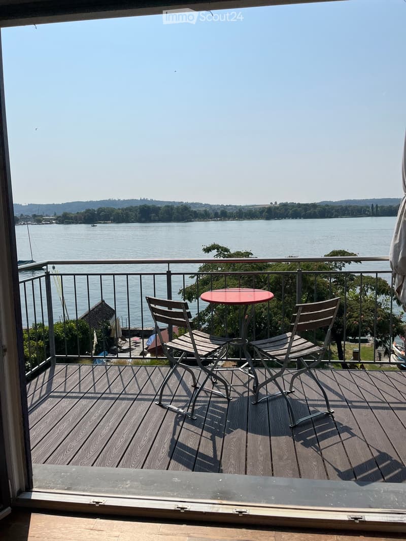 Balcony with wooden decking, metal railing, and a red round table with two wooden chairs, overlooking a lake.