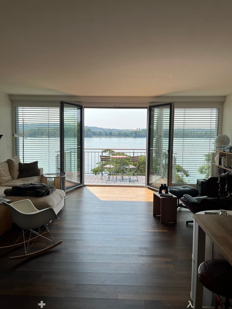 Modern living room with large windows offering a scenic view, featuring a sofa, rocking chair, small table, and a balcony with outdoor furniture.