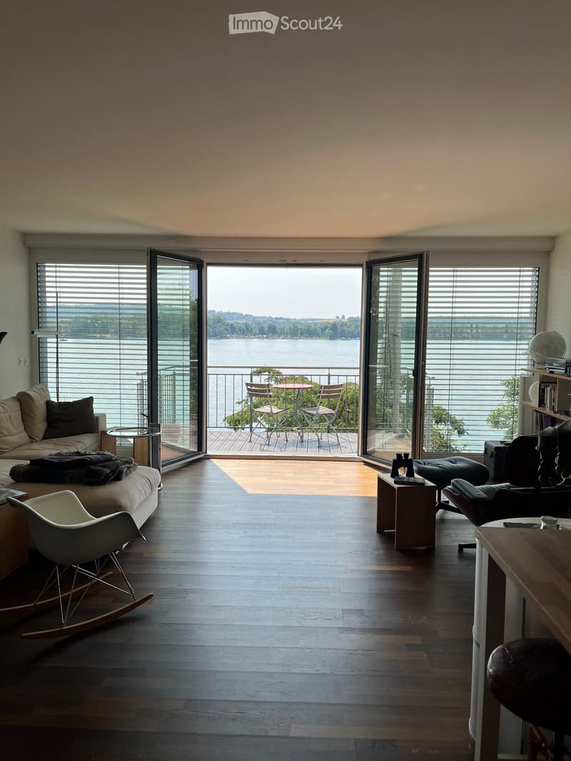 Modern living room with large windows offering a scenic view, featuring a sofa, rocking chair, small table, and a balcony with outdoor furniture.