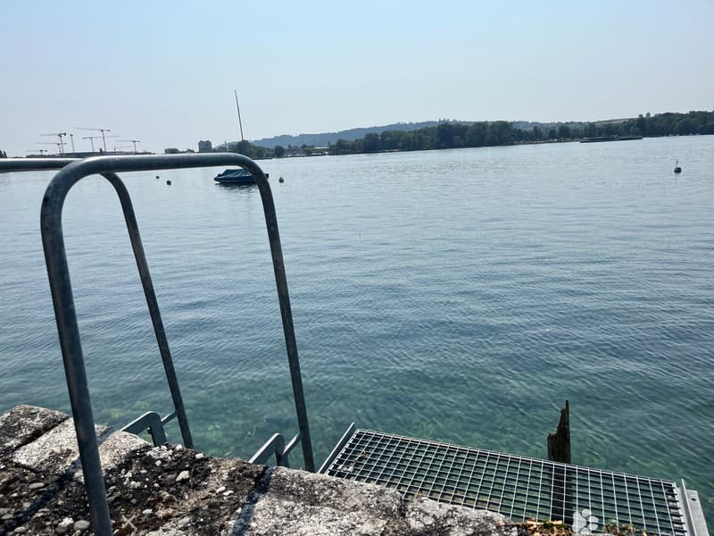 View of a calm lake with a small boat, distant construction cranes, and a distant shoreline with trees and buildings.