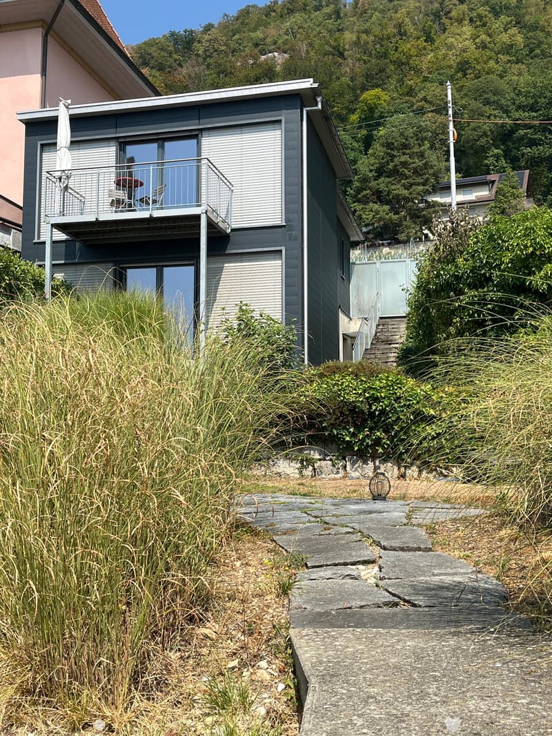 Modern two-story house with grey siding, large windows, and a balcony overlooking a scenic hillside.