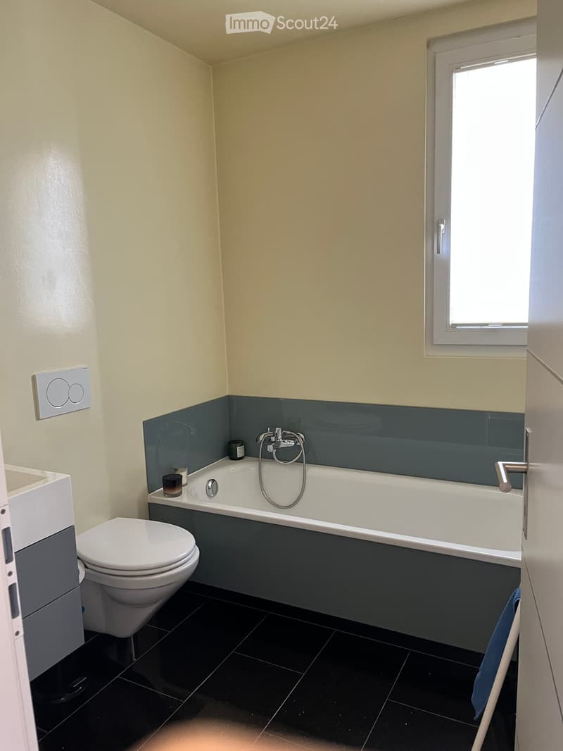 Modern bathroom with a bathtub, toilet, sink, and a small window. The walls are painted in a light beige color with a dark blue accent around the bathtub area. The floor is covered with black tiles.