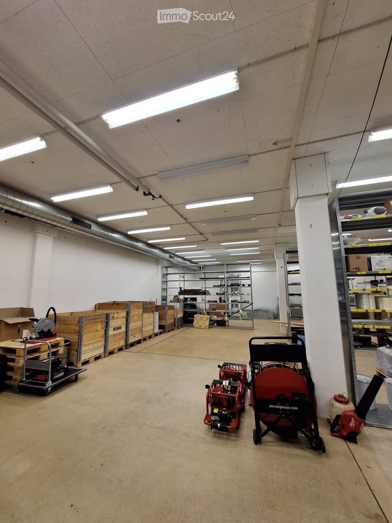 Empty warehouse space with concrete floors, white walls, exposed pipes, and fluorescent lighting. Contains storage shelves, wooden pallets, and various equipment.