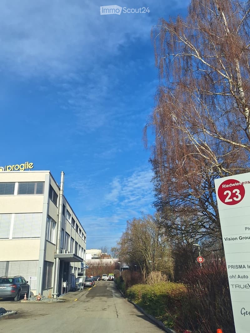Modern multi-story building with signage for "Progile" and "Riedwiesen23" visible, surrounded by trees and parked cars.