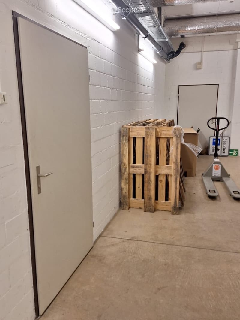 Empty storage area with concrete floor, white brick walls, and a wooden pallet stack. A door is visible on the left, and a forklift is positioned near the back wall.