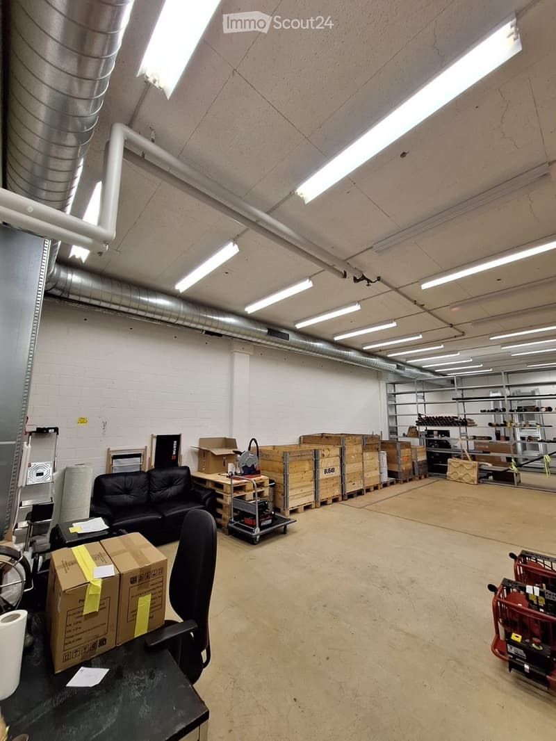 Large industrial-style room with exposed pipes and lighting, containing a black sofa, office chair, table, shelves, and storage boxes.