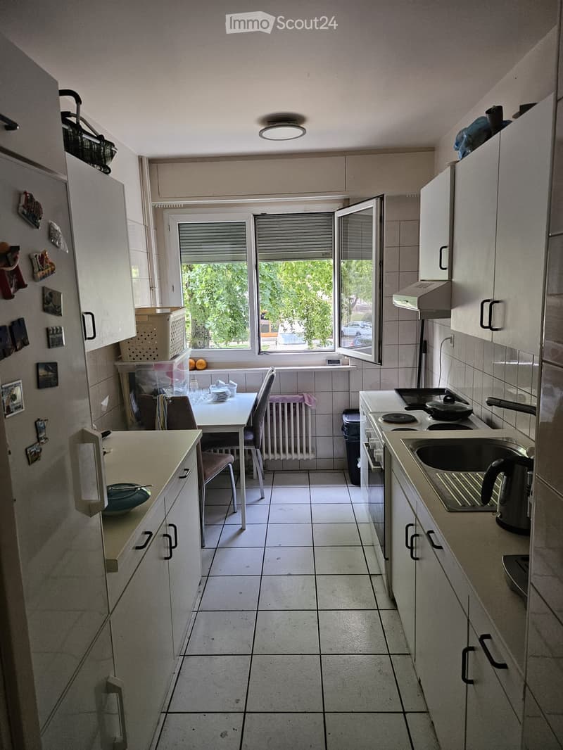 A compact kitchen with white tiled floors, white cabinets, a sink, stove, and a small dining area with a table and chairs. The kitchen has a window with blinds and a view of greenery outside.