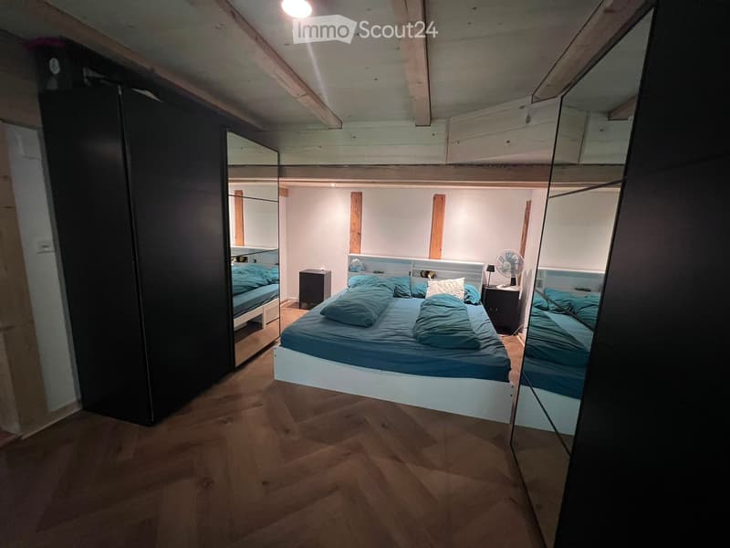 Cozy bedroom with a single bed, nightstands, a fan, and a large mirror. The room features wooden flooring and exposed wooden beams on the ceiling.