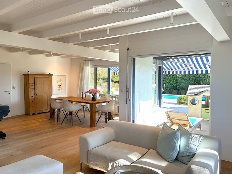 Bright living and dining area with wooden flooring, white sofa, coffee table, dining table with chairs, and a view to a patio with a swimming pool.