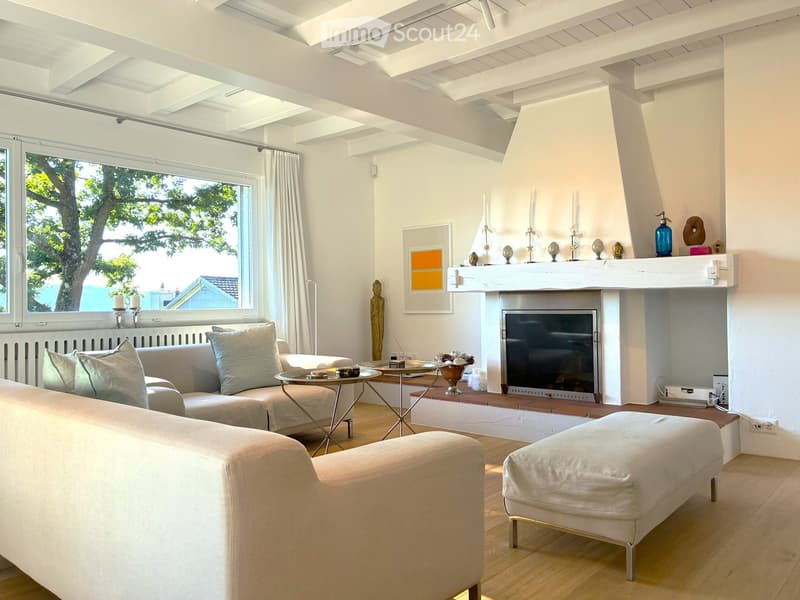 Bright living room with white walls, wooden floor, large window with view, fireplace, sofa, ottoman, coffee table, decorative items on mantel.