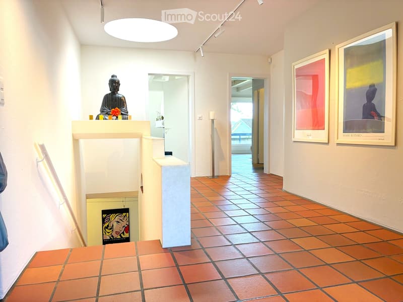 A modern hallway with terracotta tiled flooring, white walls, and framed artwork. Features a small altar with a Buddha statue and candles, and a recessed ceiling light.