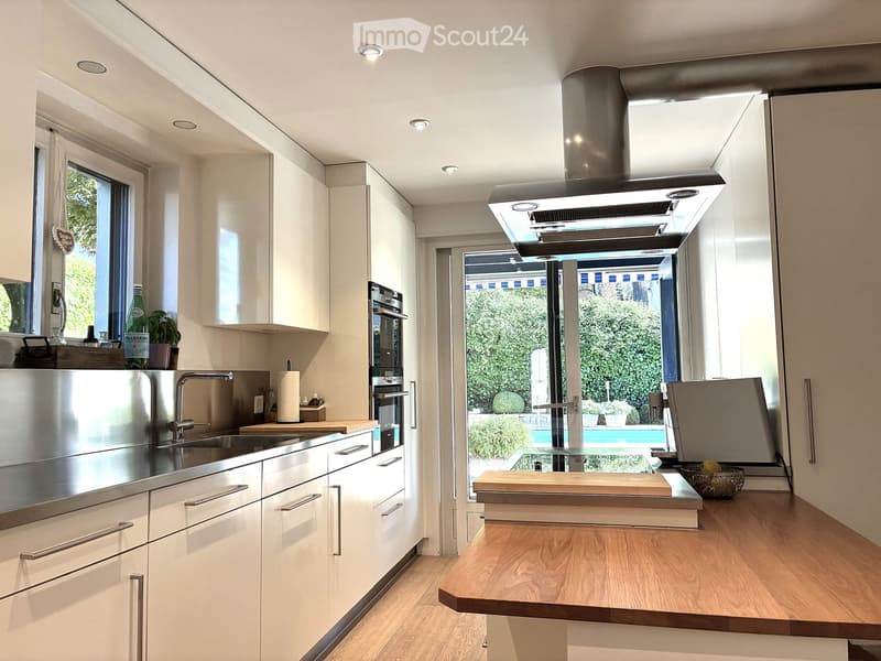 Modern kitchen with white cabinets, stainless steel appliances, island countertop, and large window overlooking a garden and pool.