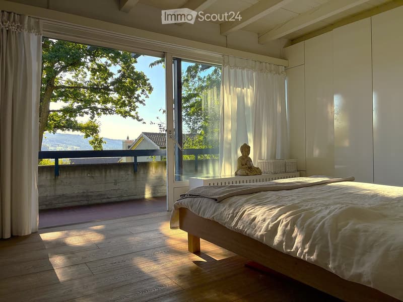 Bright bedroom with wooden floor, large window and balcony overlooking a scenic view, featuring a bed, a sitting statue, and wicker baskets.