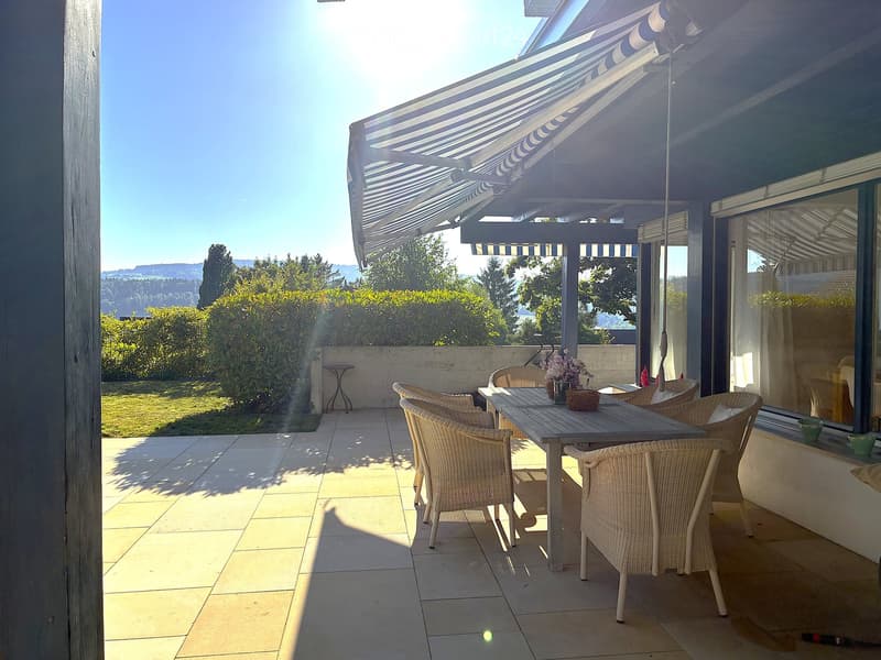 A covered outdoor patio with a dining table and chairs, overlooking a garden and distant hills.