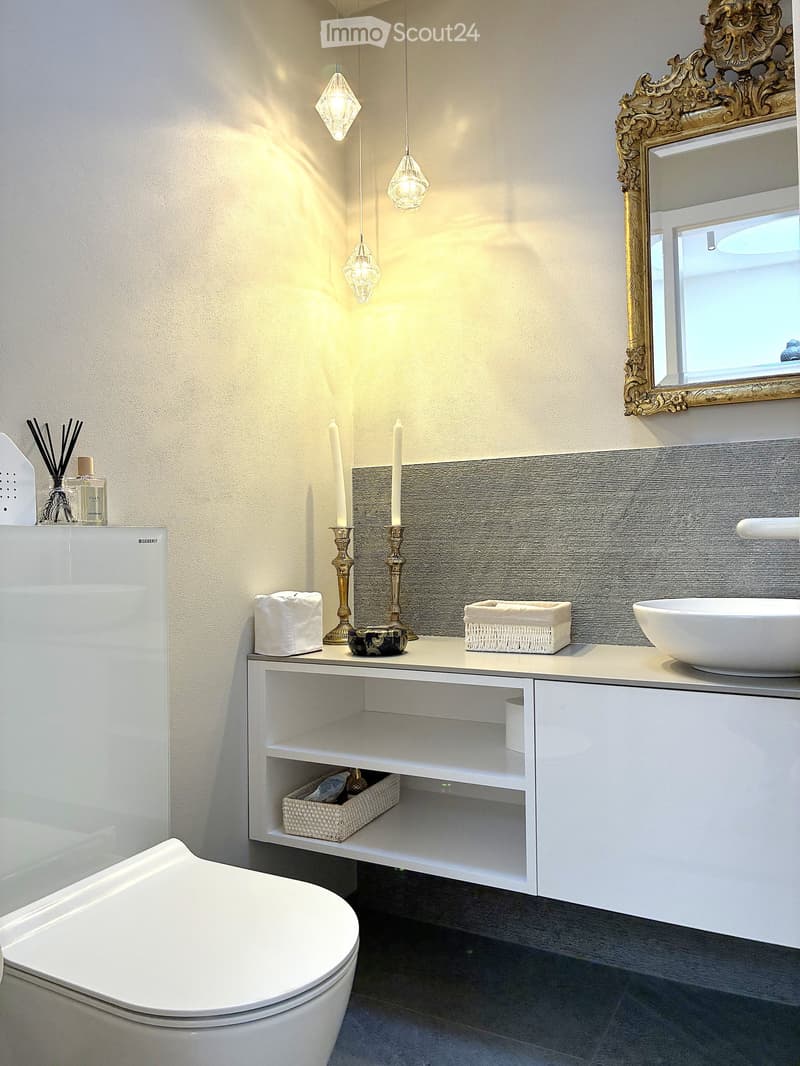 Modern bathroom with white sink, toilet, and vanity cabinet. Features a large ornate mirror, candles, and decorative items on the counter.