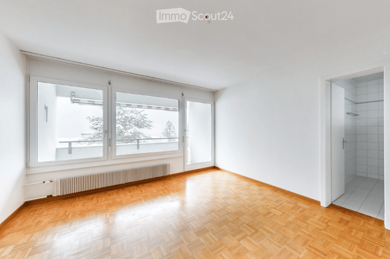 Empty room with wooden flooring, large windows, radiator, and a view of a balcony area.