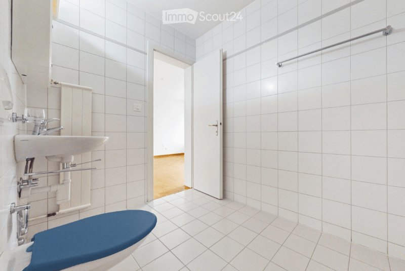 Modern bathroom with white tiled walls and floor, a blue toilet seat, a white sink with chrome faucet, and a door leading to another room.